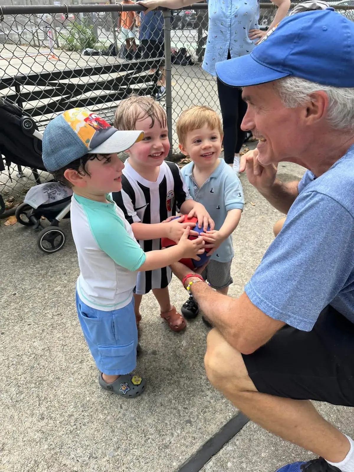 Coach Mike crouching at eye level with three young boys, all holding a ball together and laughing